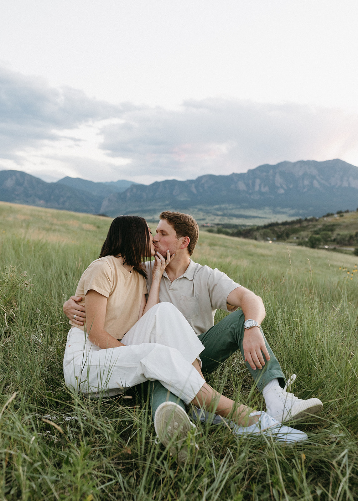 A couple in neutral attire shares a kiss in front of The Flatirons during their engagement session in Boulder