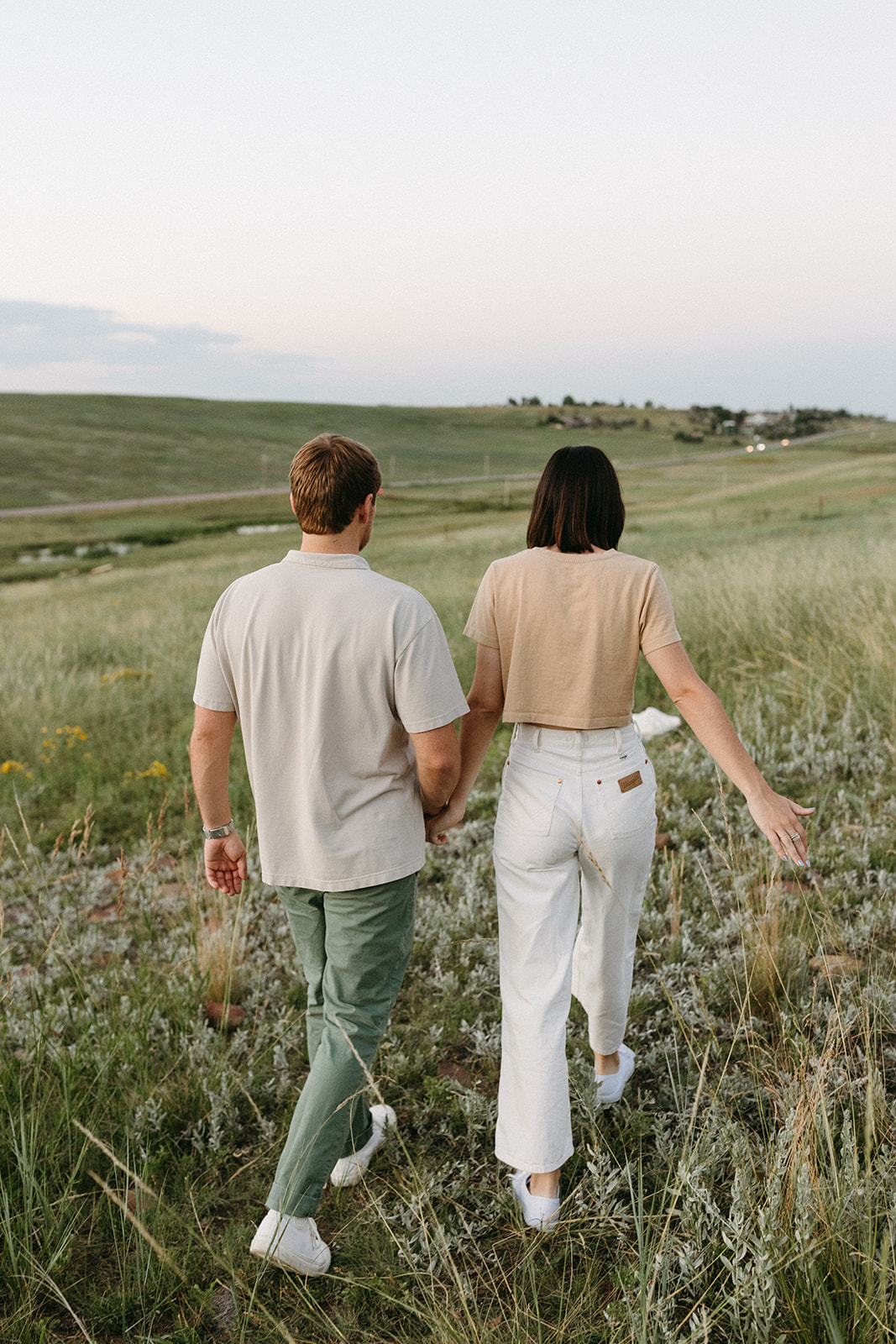 A woman in a beige cropped tee and white jeans walks and holds her fiancé's hand during a wildflower engagement session