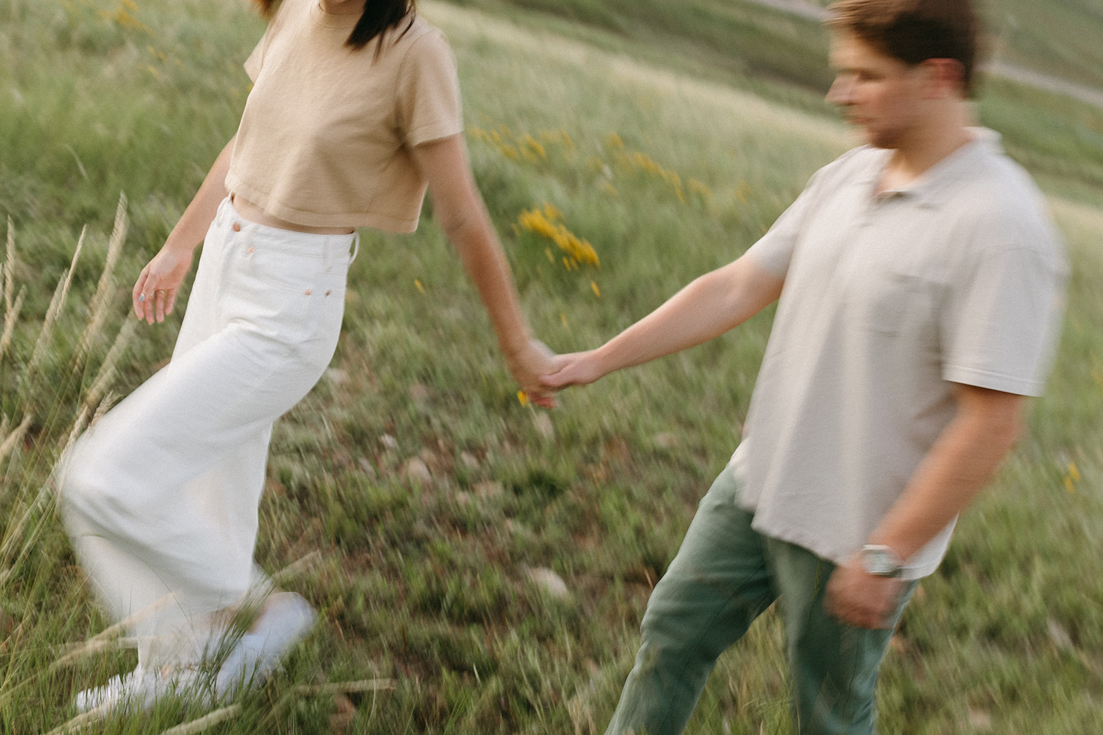 A man in a taupe polo shirt and forest green chino pants holds his fiancée's hand during a wildflower engagement session