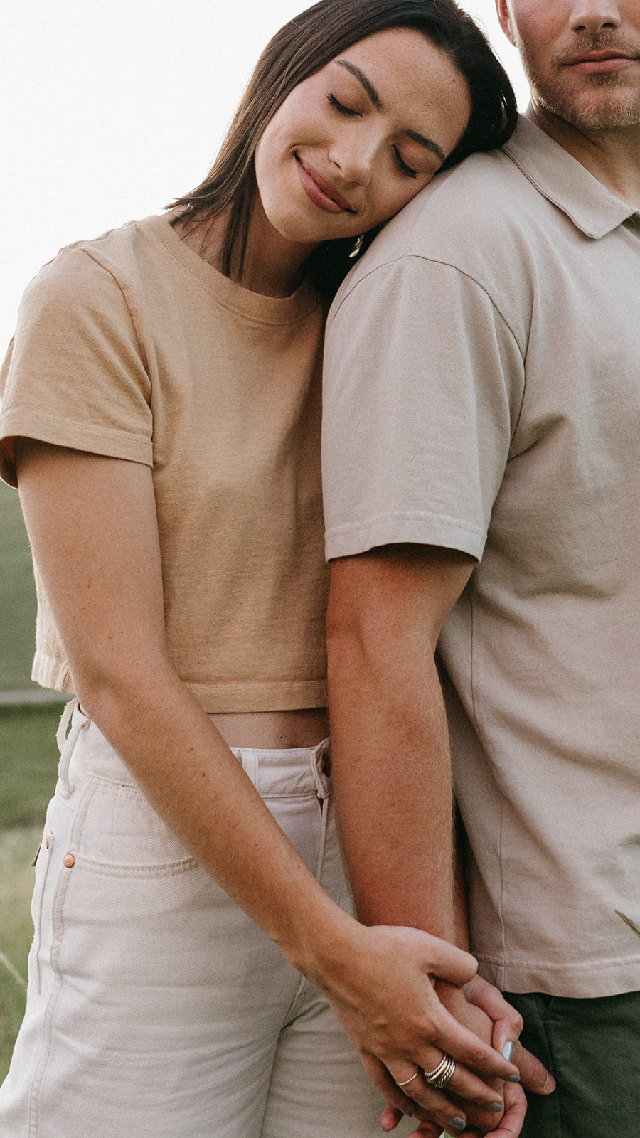 A woman in a beige cropped tee and white jeans leans her head on her fiancé's shoulder during their editorial engagement