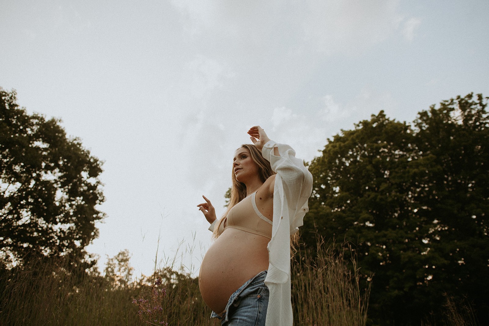Pregnant woman poses in an oversize button up and jeans for her sunset maternity photoshoot