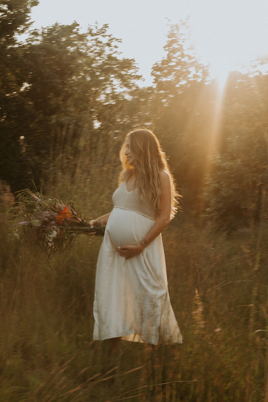 Woman with long hair wearing a white dress cradles her growing belly during a sunset photosession 