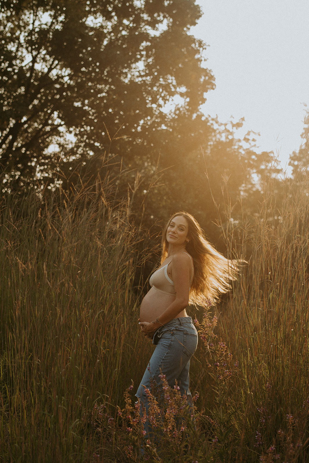 Expecting mom in jeans and a white bra flips her hair as the sun sets behind her 