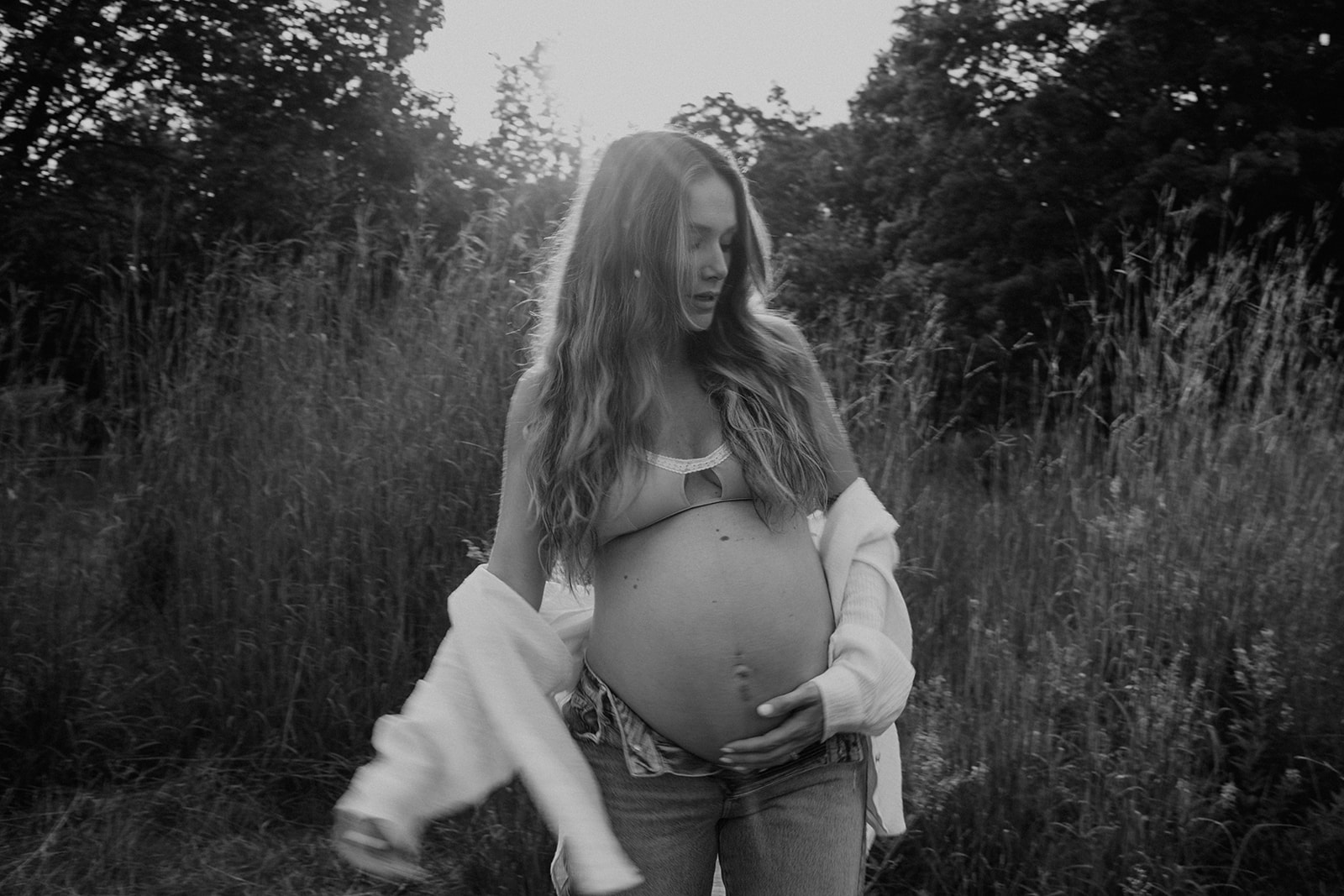 Pregnant woman embraces her belly and poses her for maternity photographer in an oversize button-up shirt