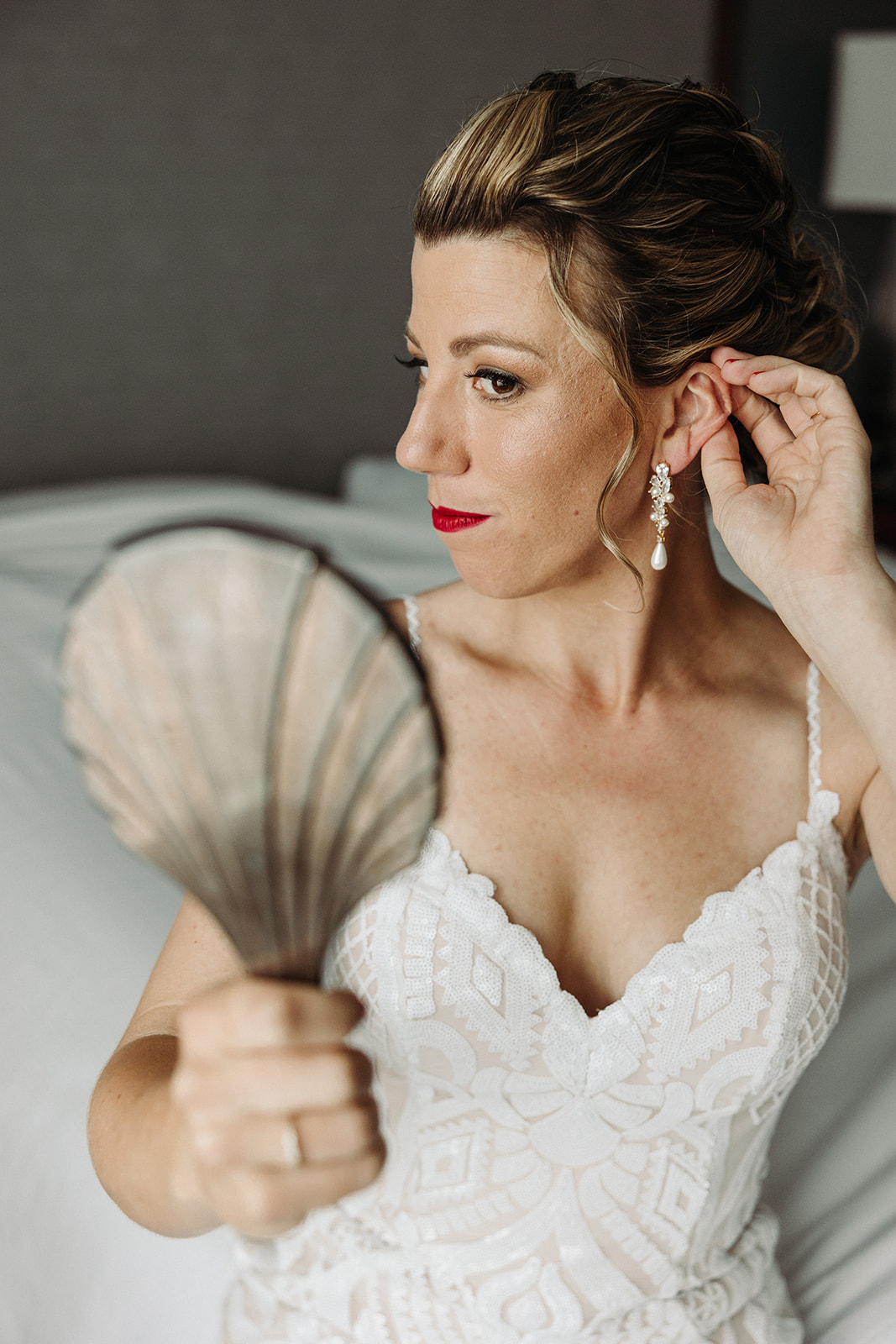 A bride sits on a bed in her suite while using the mirror to adjust the hair around her ear.