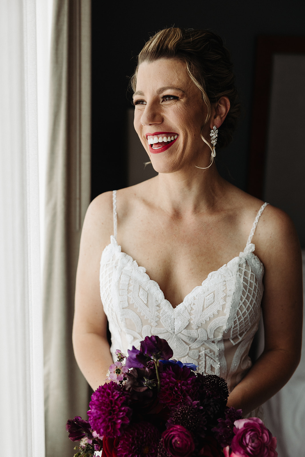 A beaming bride smiles widely as she holds her vibrant purple bouquet in the natural window lighting.