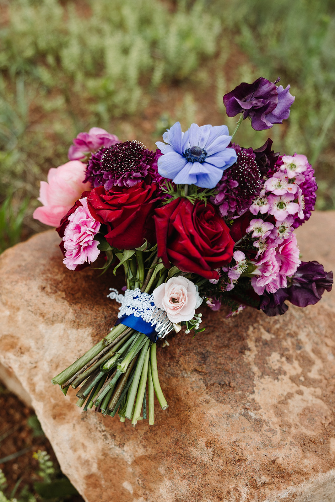 A bold bouquet with rich reds, purples, blues, and pinks. The stem is wrapped in a blue ribbon with personal touches.