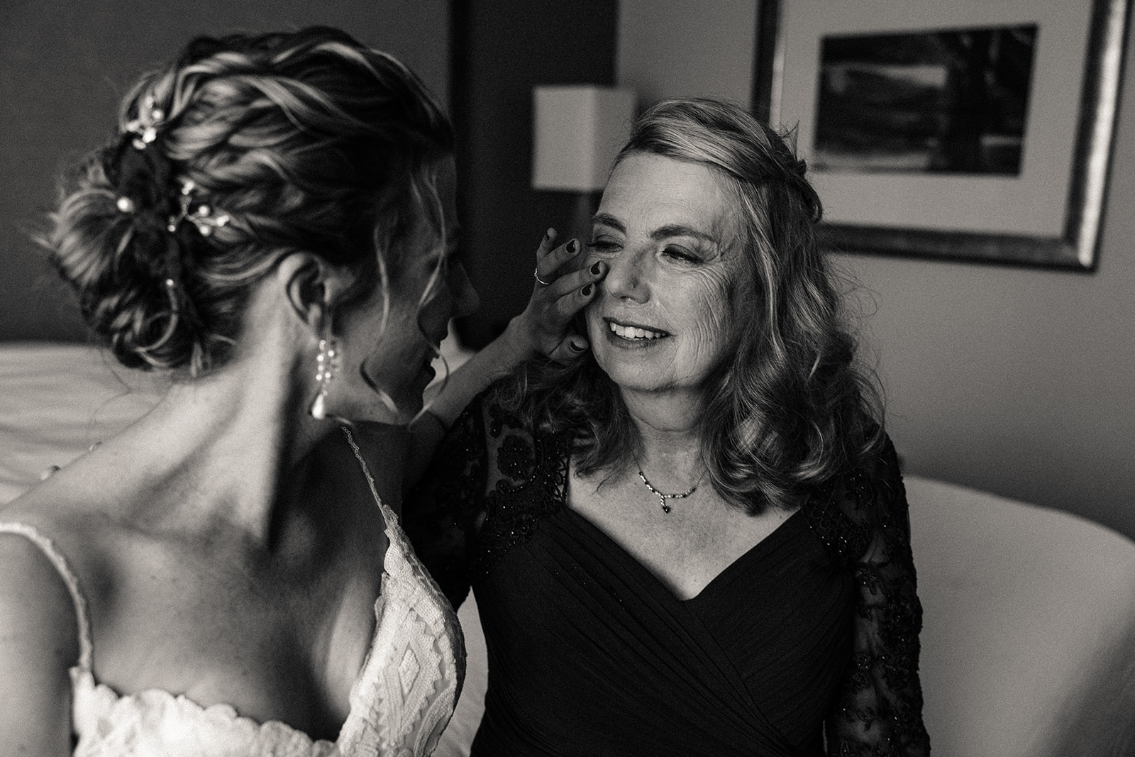 Bride wipes her mother's face as they smile and share tears of joy on the bed in the suite.