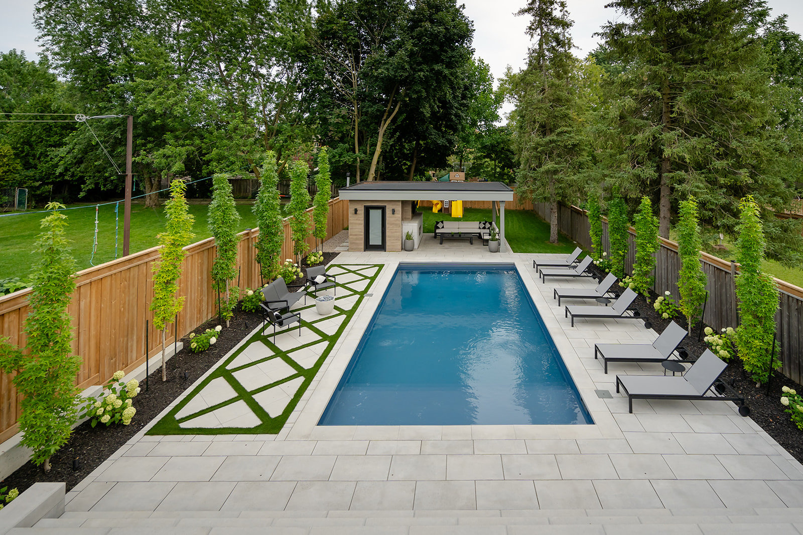 An inground pool with lounge chairs on the right and 3 chairs on the left.  Along the fences are trees planted.