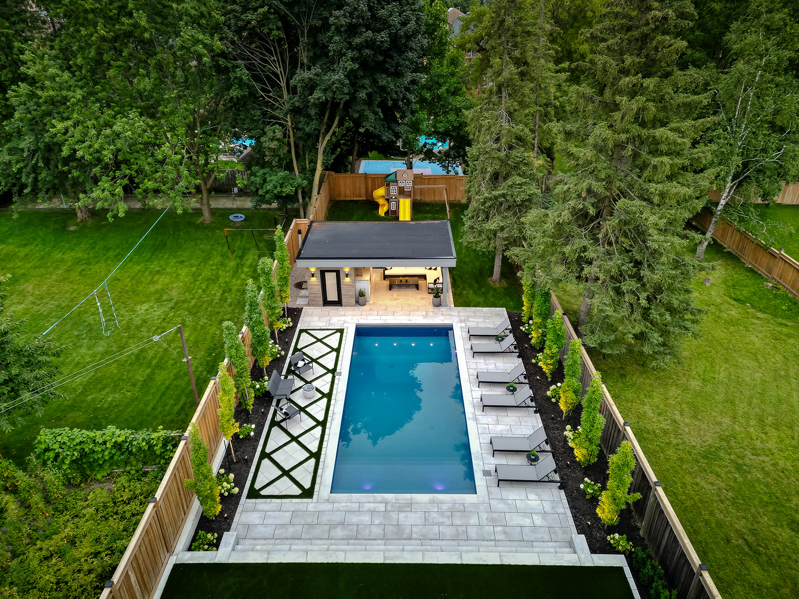 An inground pool with lounge chairs on the right and 3 chairs on the left.  Along the fences are trees planted.
