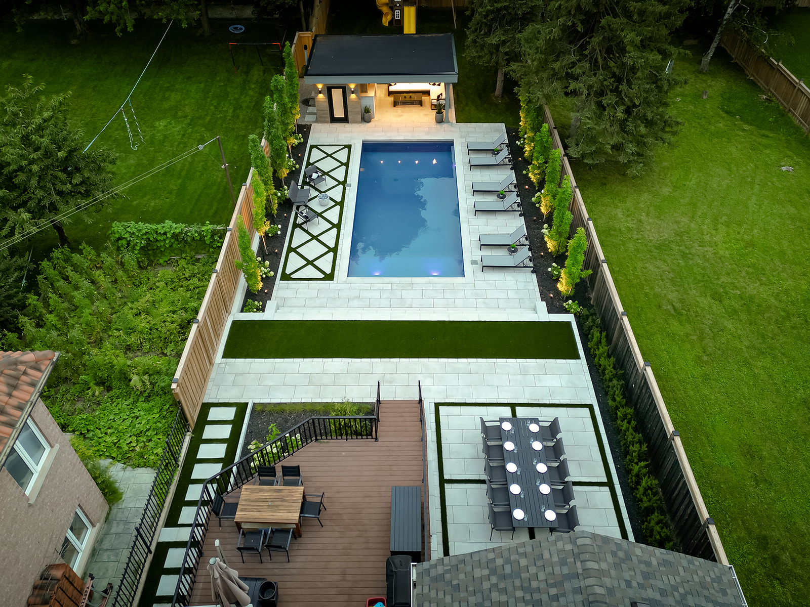 An inground pool with seating on either side, a pool house above and seating on the deck and on the patio stones.