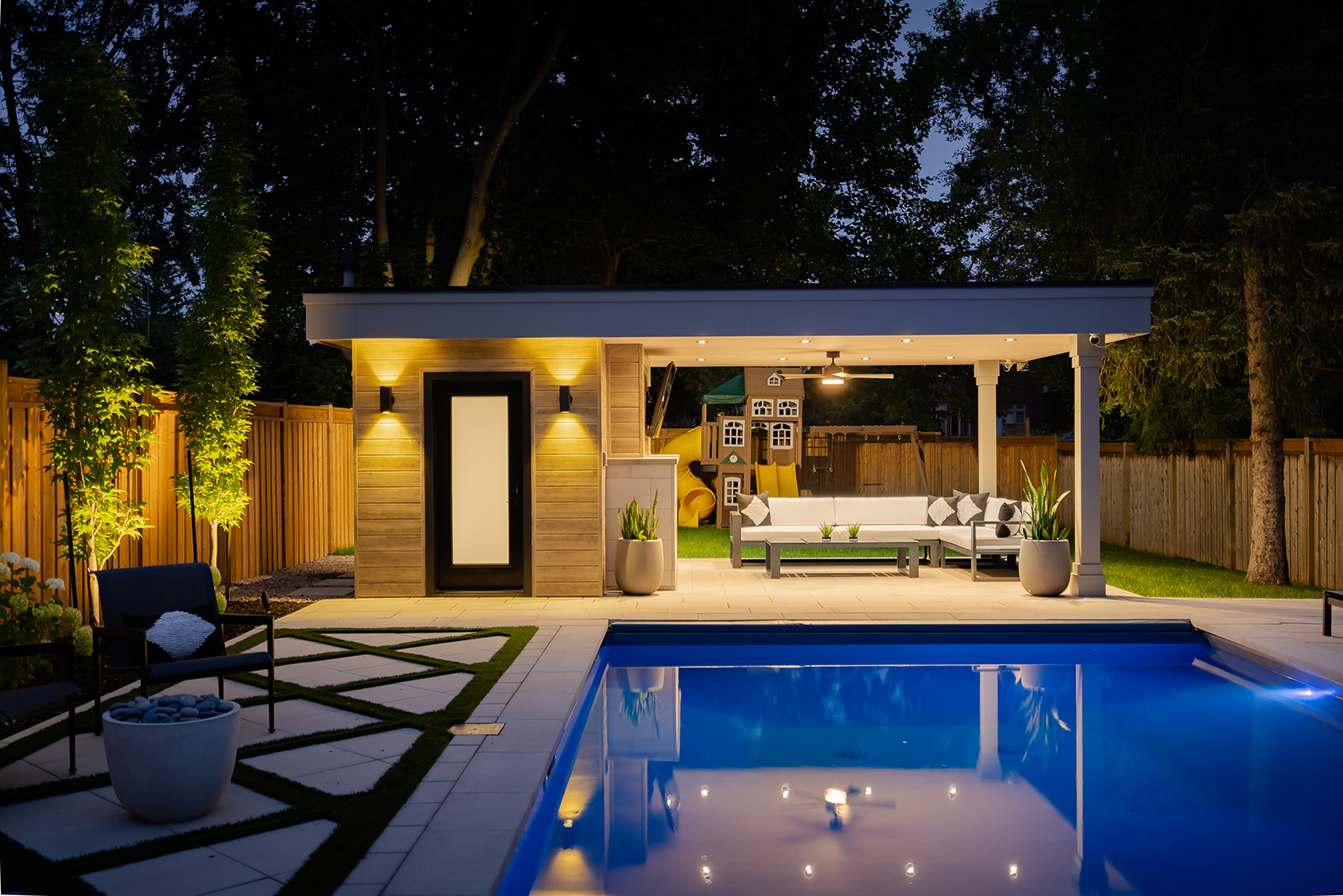 A a pool house with lights on, an outdoor patio set to the right and chairs on the left on the patio stones.