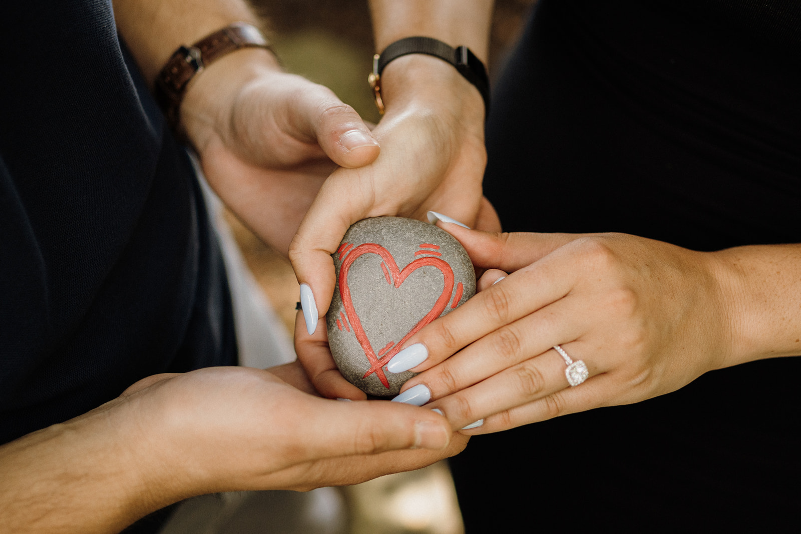 Hands holding a rock with a heart on it.  One of the hands have an engagement ring on.