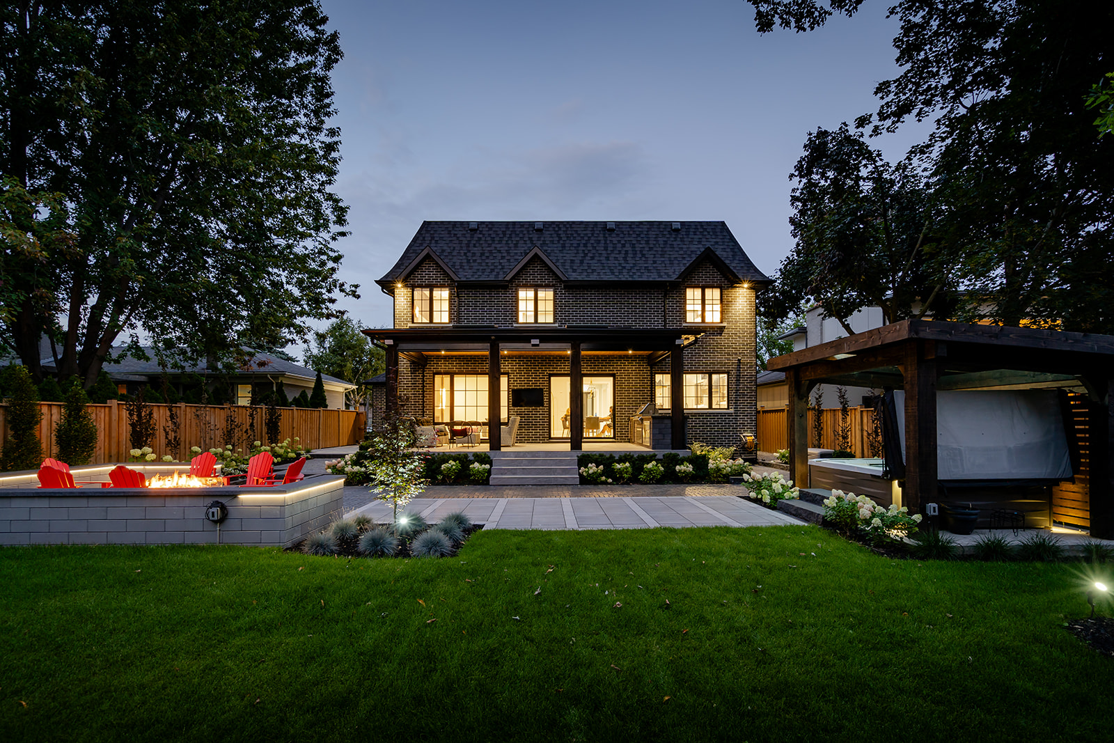 A patio setup with Muskoka chairs on the left and a hot tub on the right with lights on in the house.