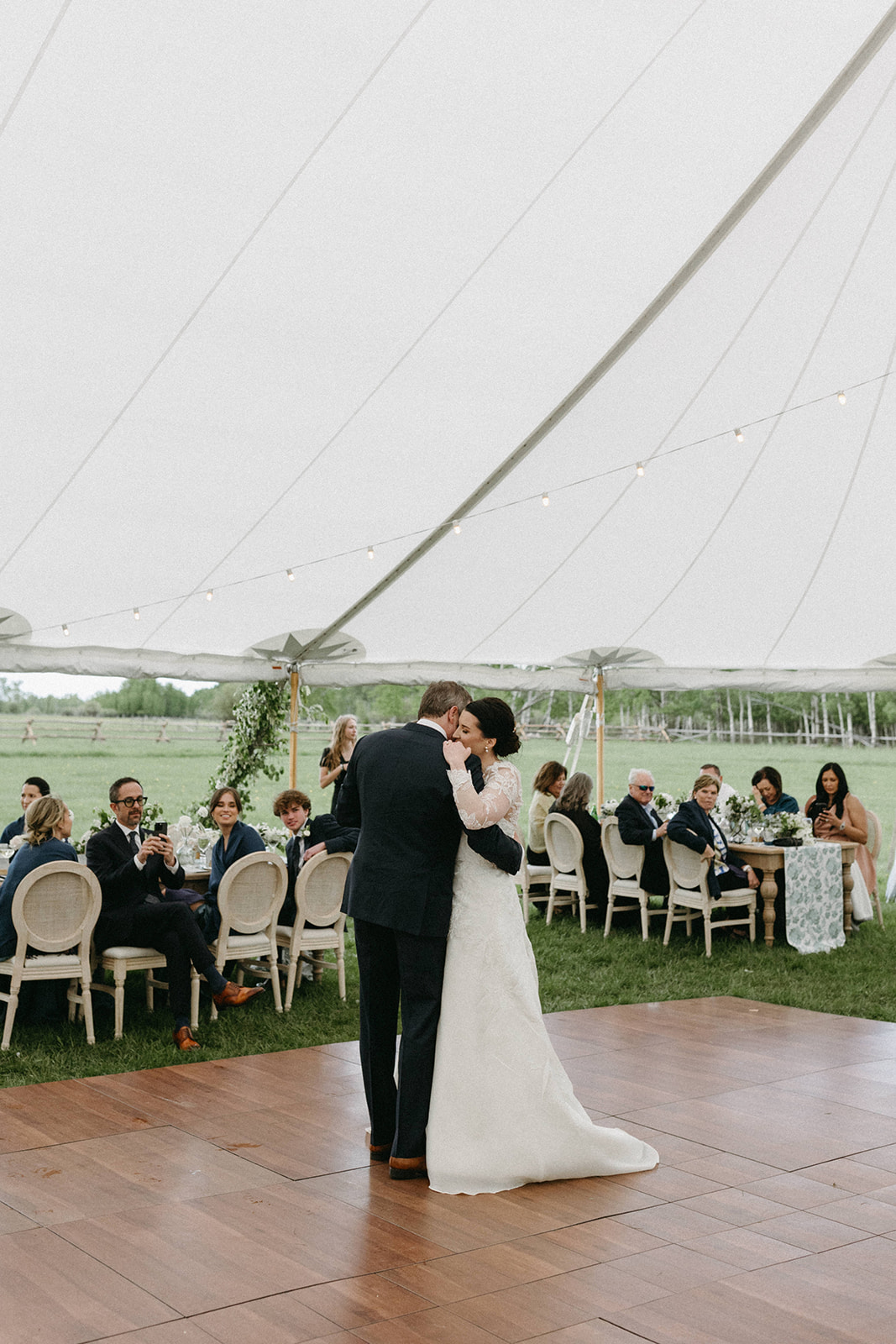 A man in a black tailored suit dances with his wife during an outdoor wedding reception in a white tent. 