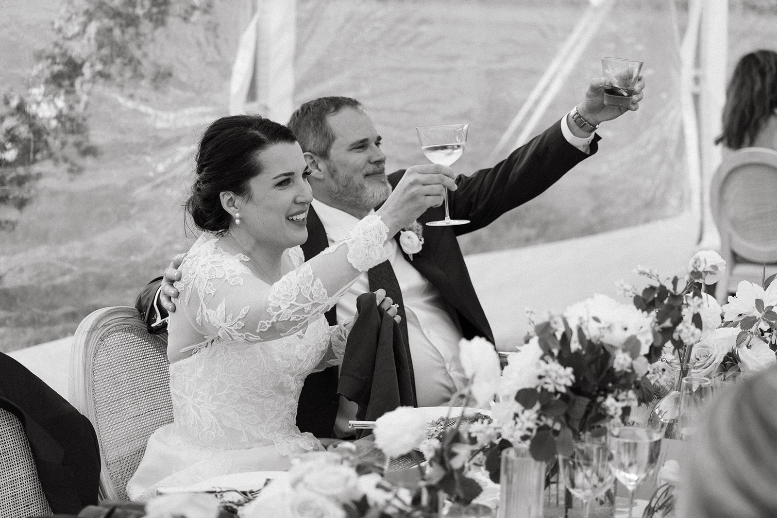 A bride and groom in elegant wedding attire hold up champagne glasses during their wedding reception toast. 