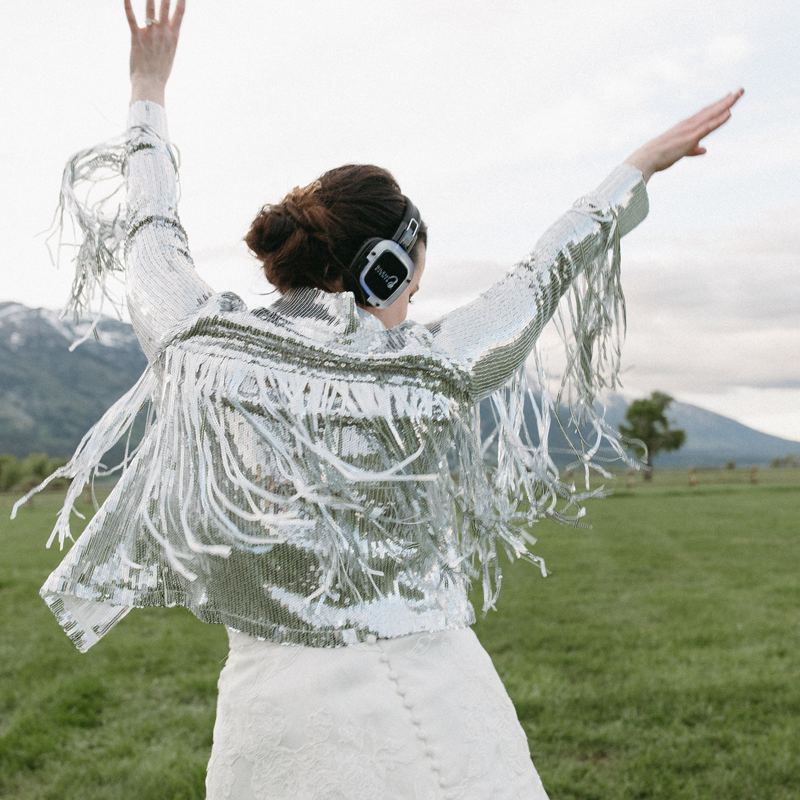 A bride dances on a ranch during a silent disco reception wearing a silver-sequin fringe jacket. 