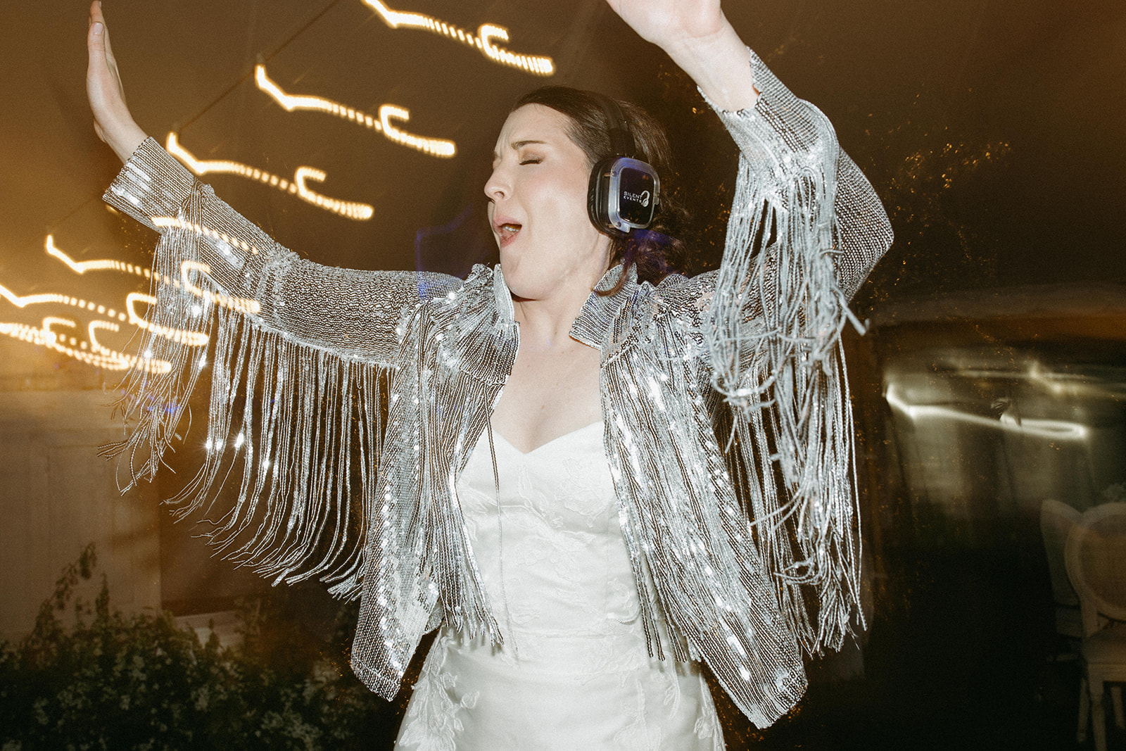 A bride dances at a silent disco wedding reception wearing a silver-sequin fringe jacket. 
