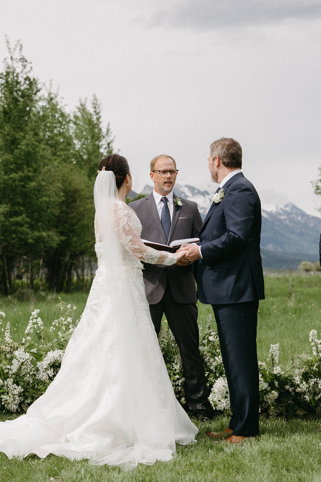 A bride in a long-sleeve, white lace wedding gown exchanges vows with a groom in a custom tailored black suit. 