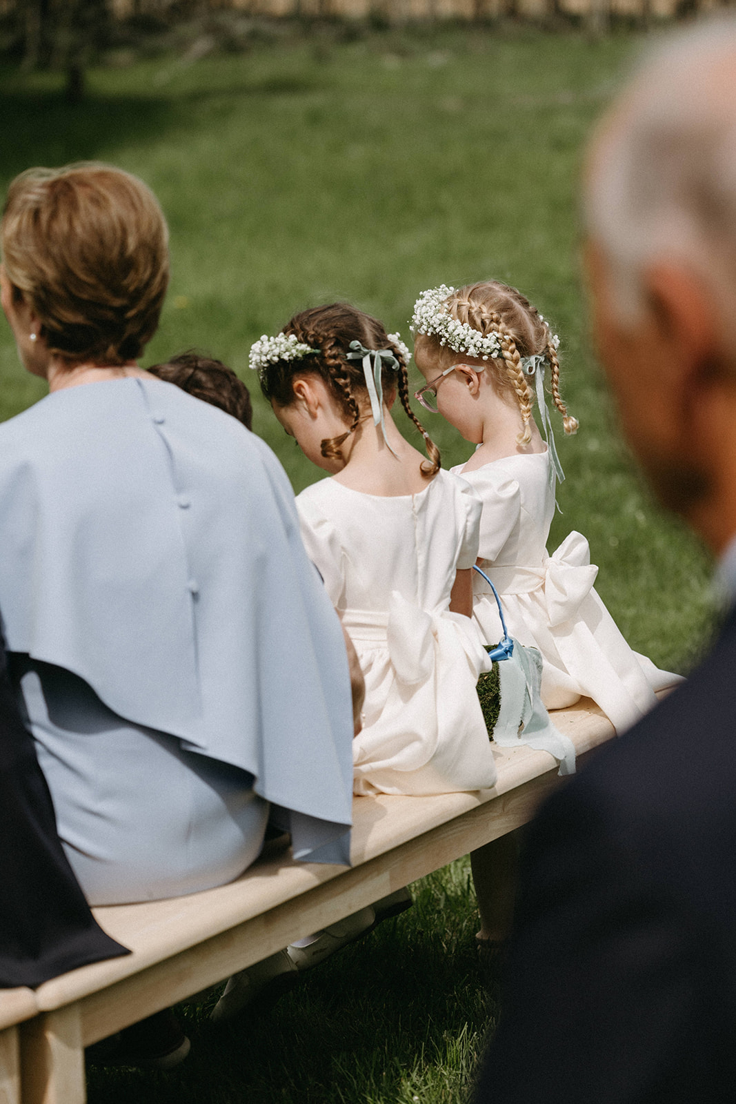 Two floral girls wearing white floral grows and white flower girl dresses sit with wedding guests on a wooden bench. 