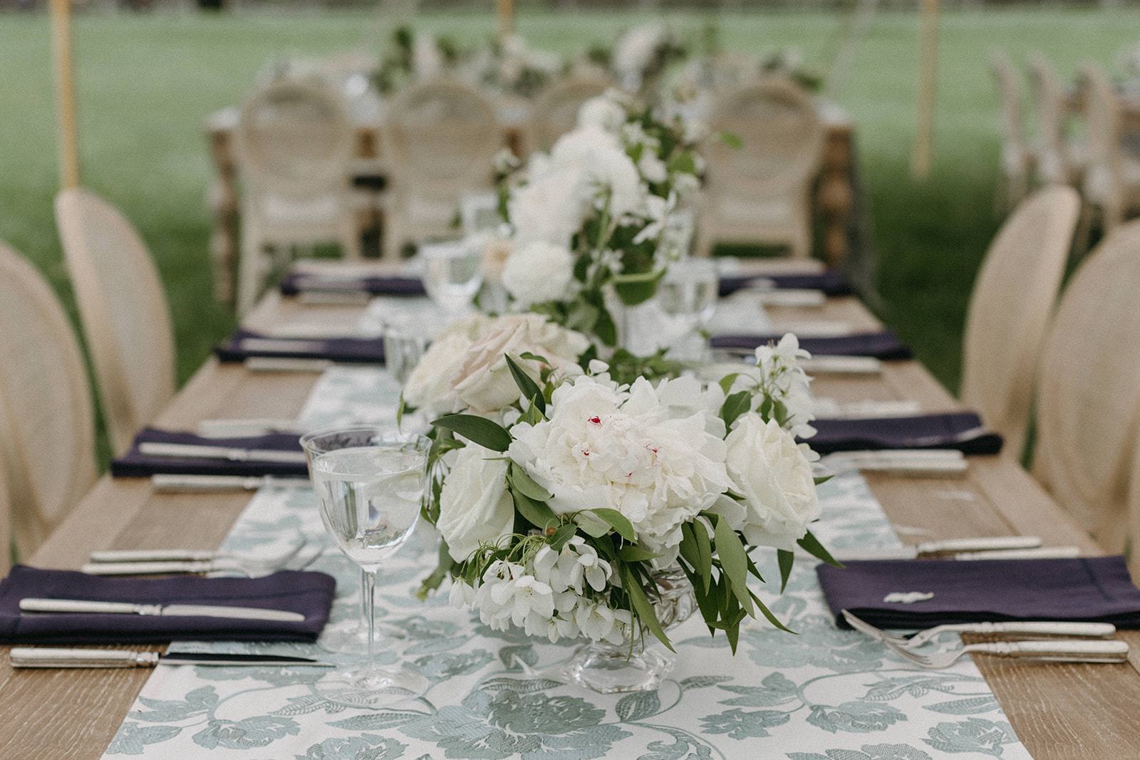An elegant wedding reception tablescape with white rose centerpieces accented with dark purple napkins.
