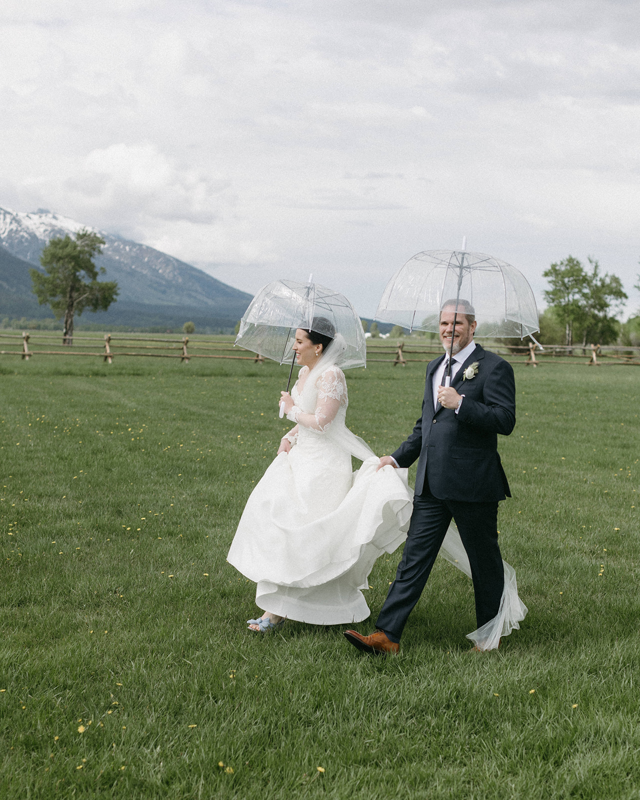 A bride and groom, holding clear bubble umbrellas, walk through a field during their wedding at Snake River Ranch. 
