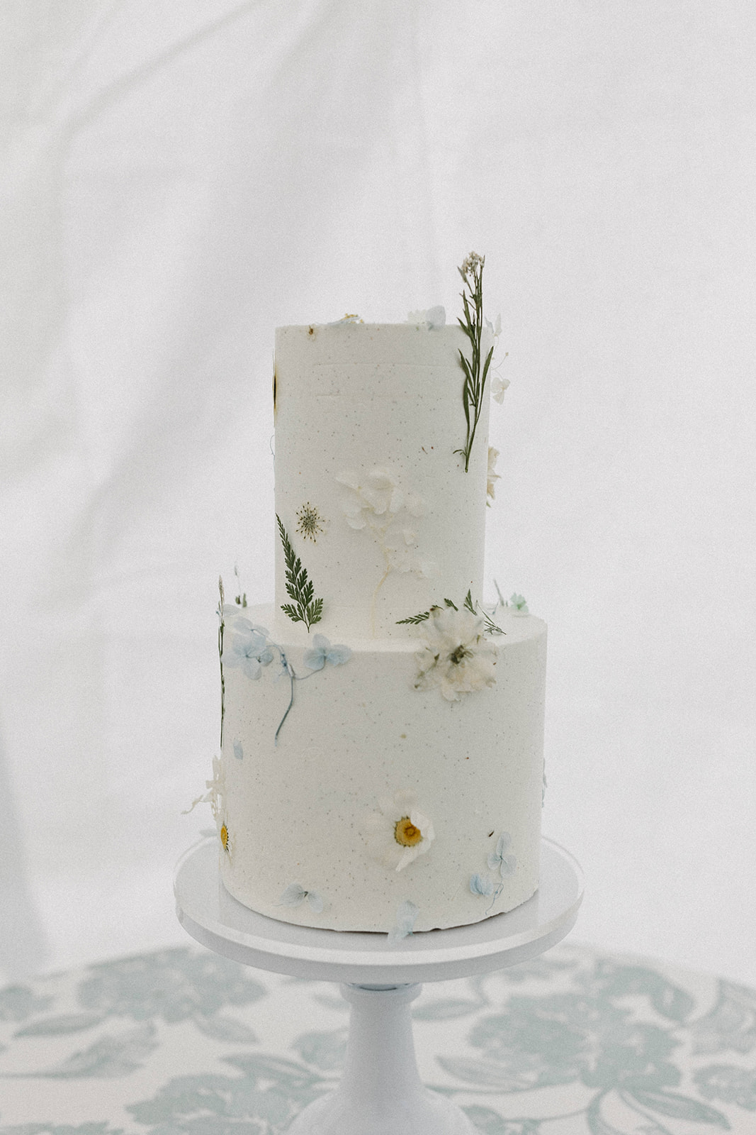 A delicate floral wedding cake with white frosting on a white cake stand. 

