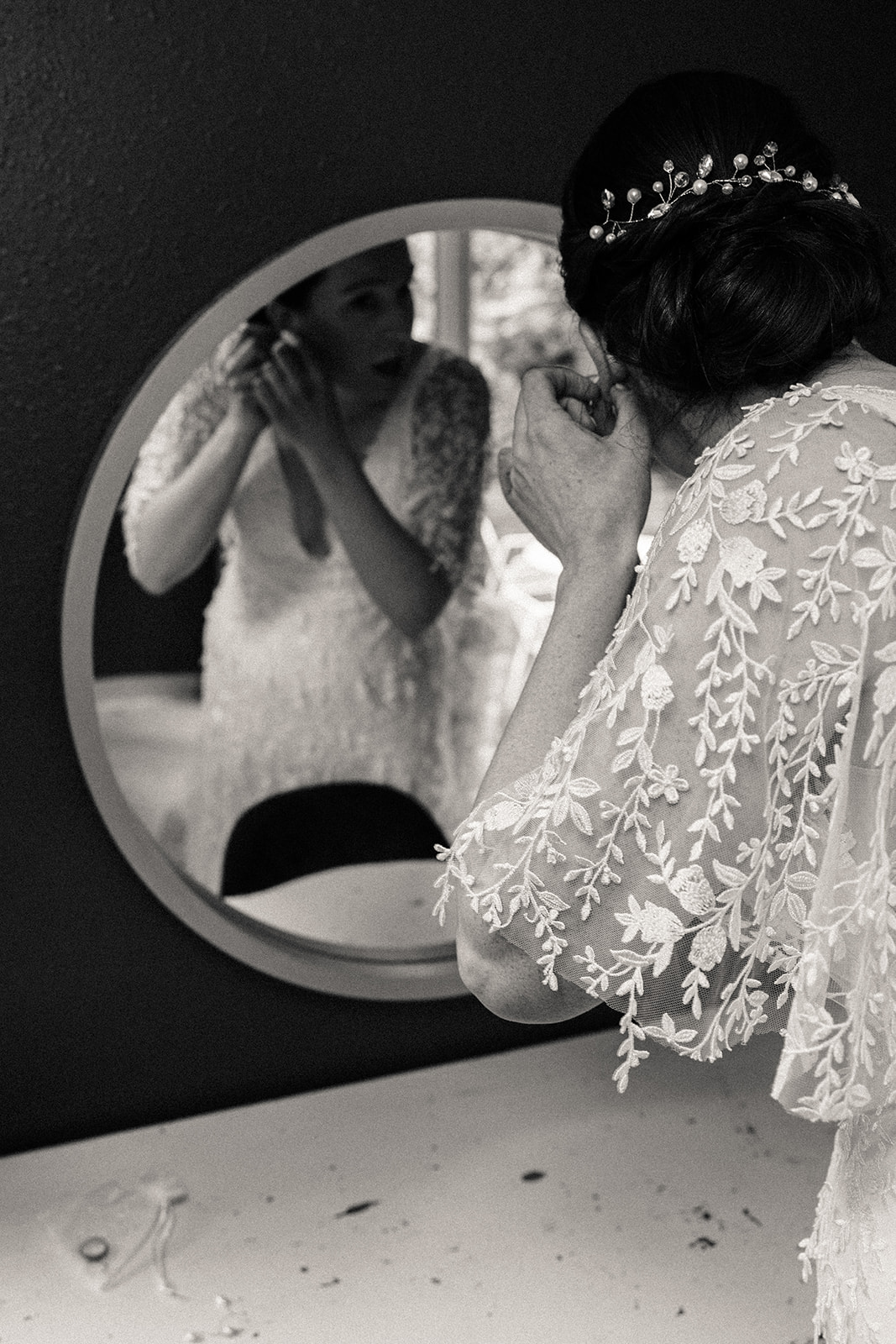 The bride bends over to put on her earrings while looking in the small, round mirror in her bridal suite.