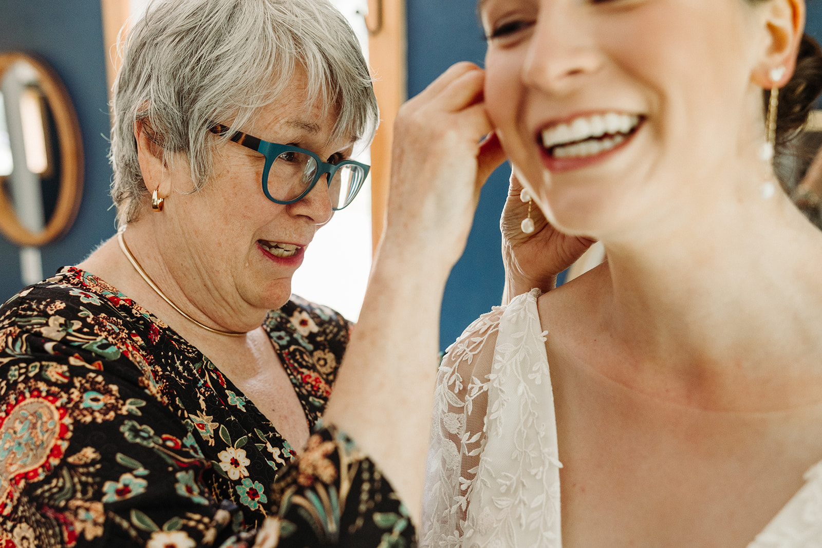 The mother of the bride puts on her daughter's dangly earrings as the bride smiles brightly.