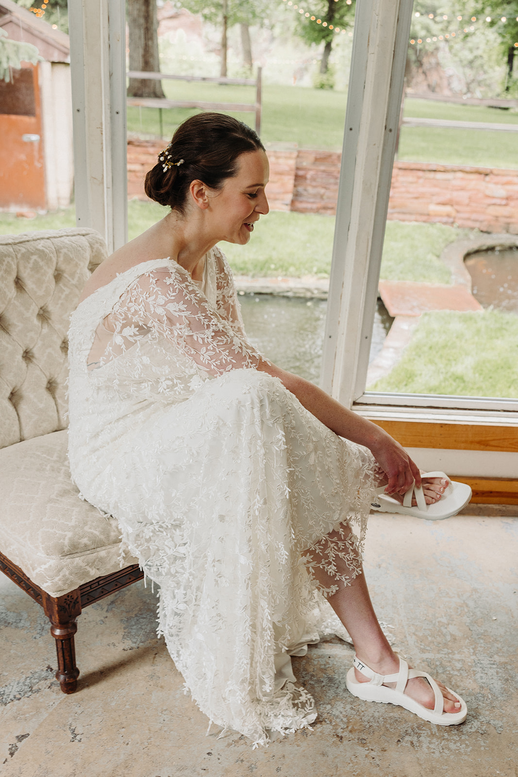 Bride in her wedding dress sits on a white chair as she puts on her white wedding chacos.