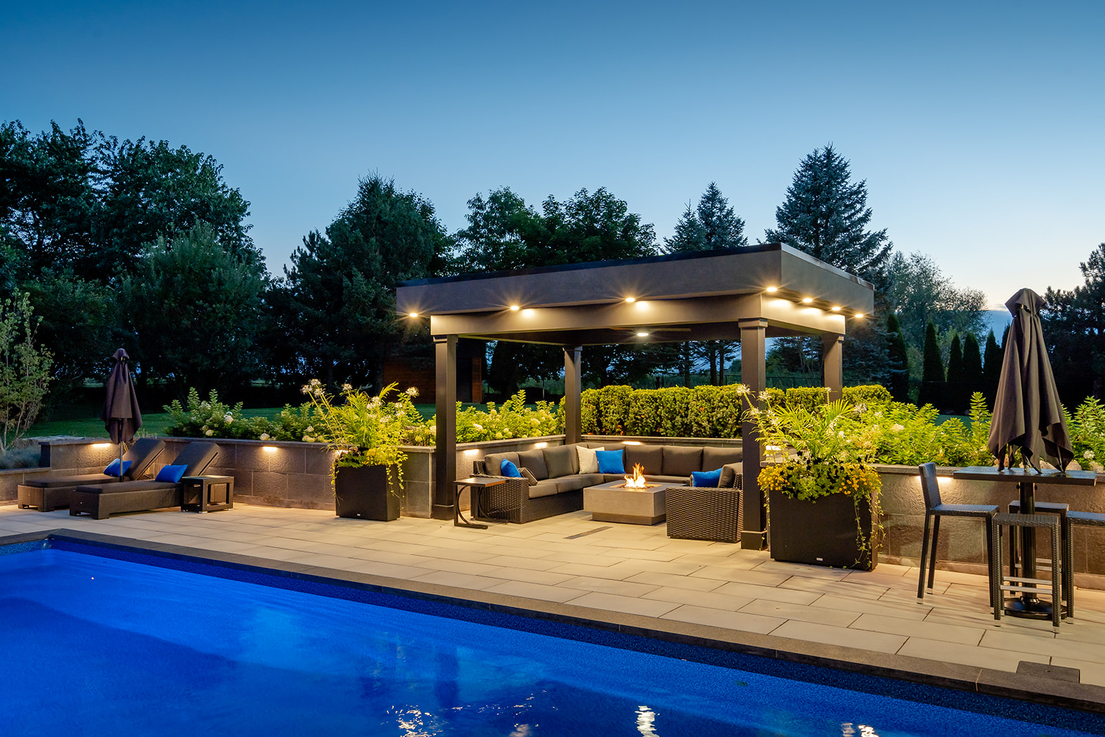 An outdoor seating area underneath a gazebo with lights turned on and the inground pool.