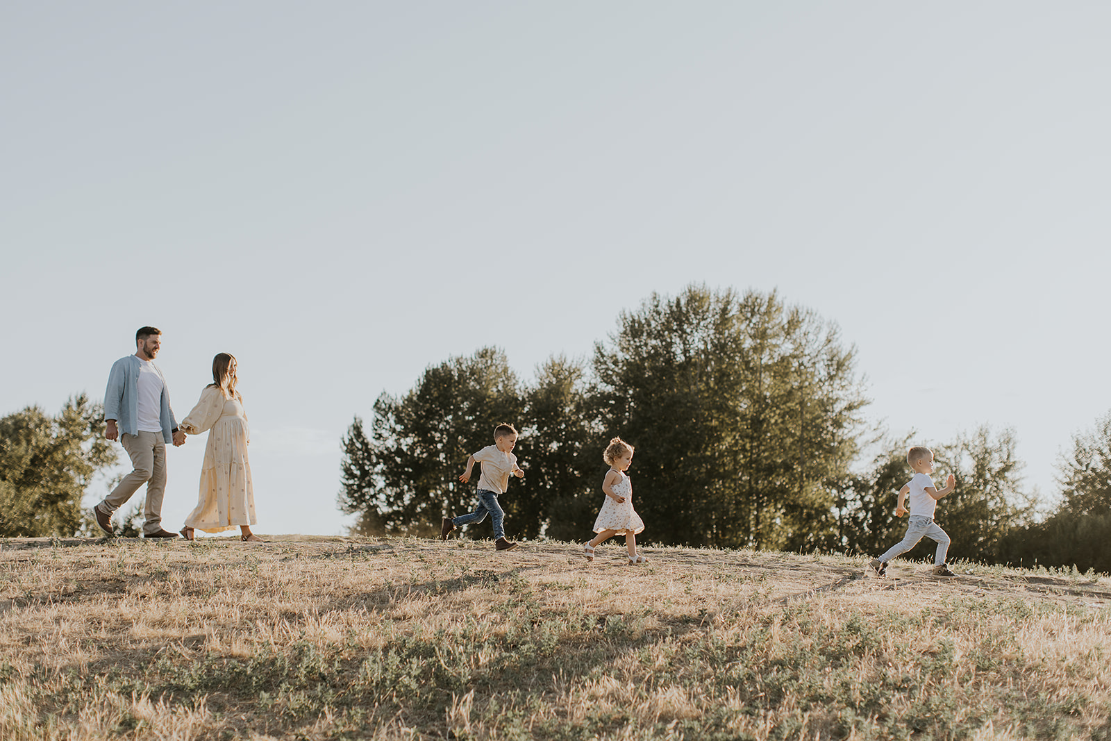 three kids running down a grassy hill with a pregnant couple following behind smiling
