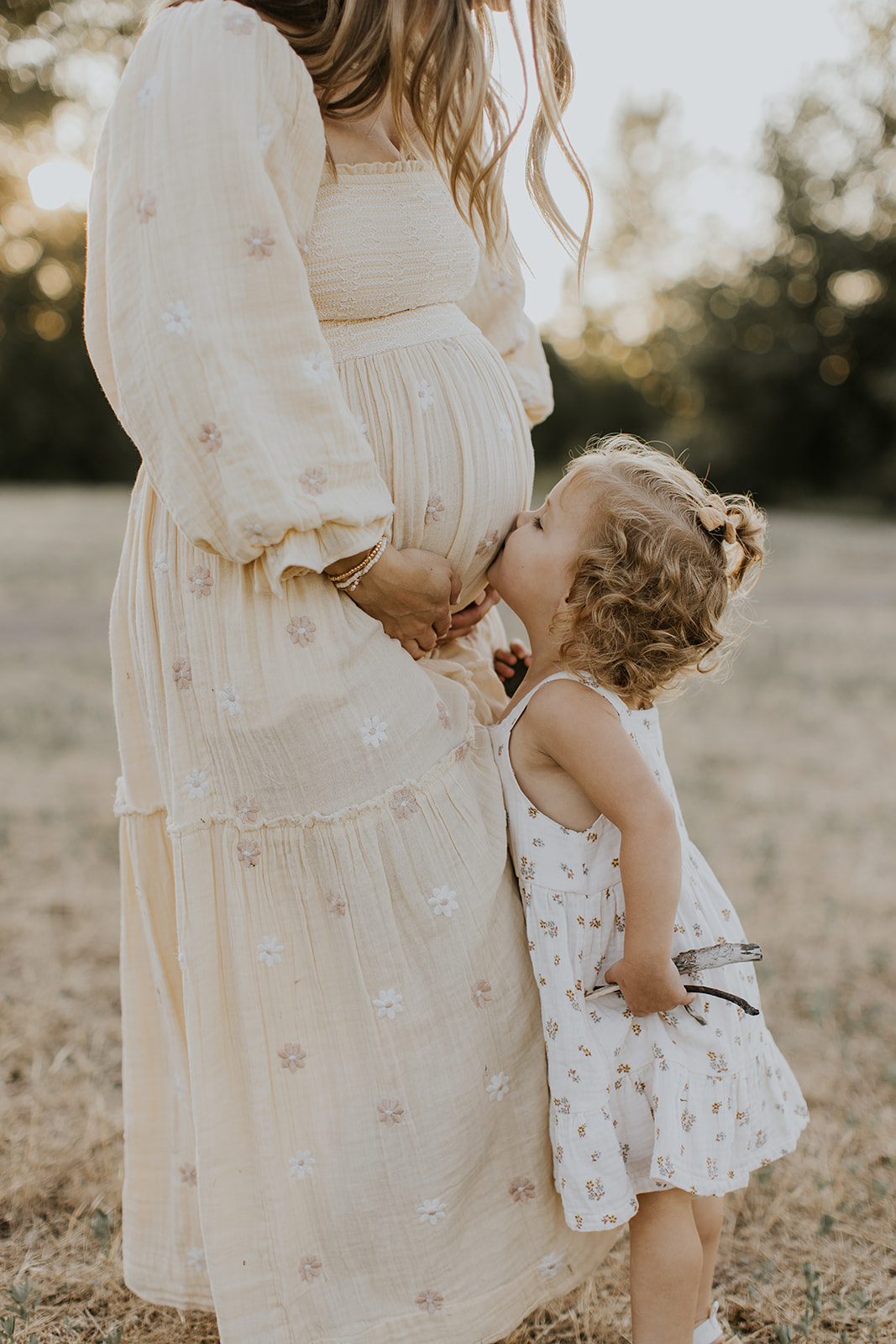 a little girl kissing her pregnant mother's baby bump while she holds flowers
