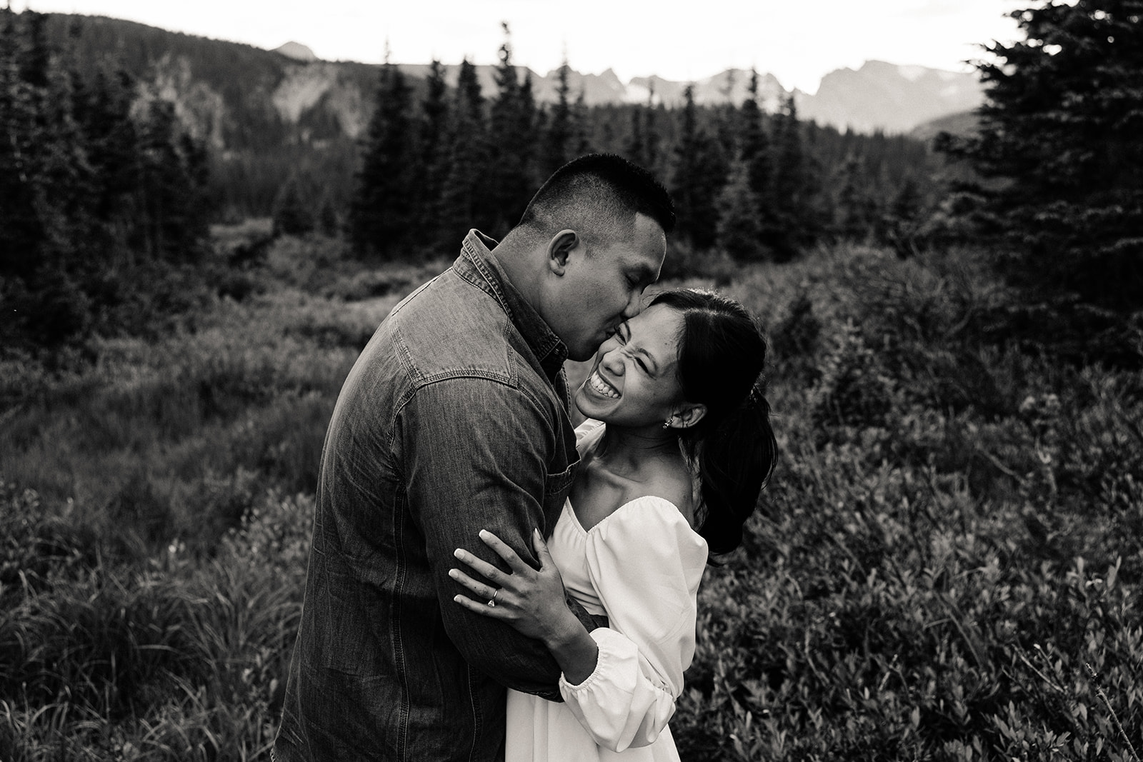 Black and white engagement photo of the pair in the tall grasses, her smiling widely as he sweetly kisses her cheek.