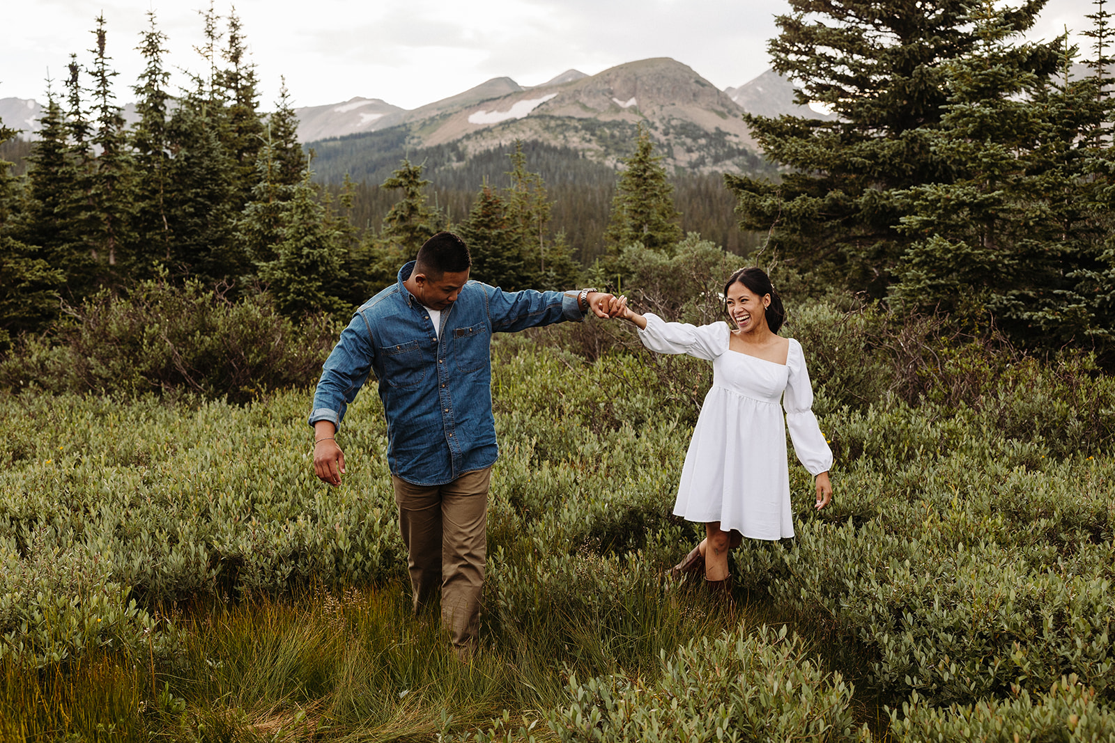 The two hold hands while walking through the meadow, with the green trees and mountains behind them.