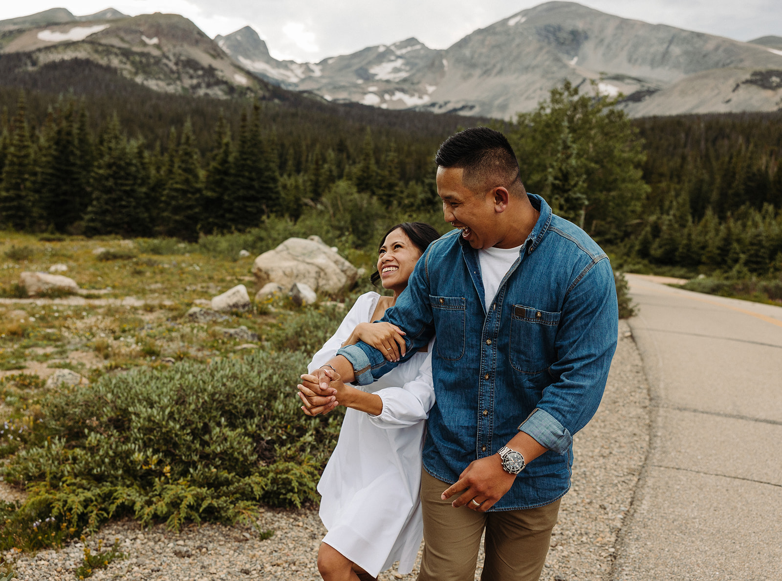 Engaged couple walk down a path in a grassy area in the mountains as the man's fiance holds his arm and smiles.