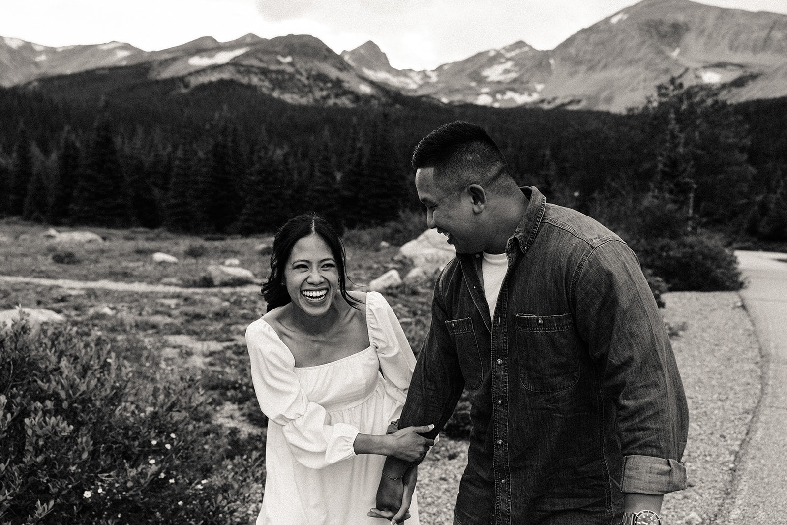 A black and white image of the engaged couple laughing as she holds his arm lower, the mountains sore behind them.