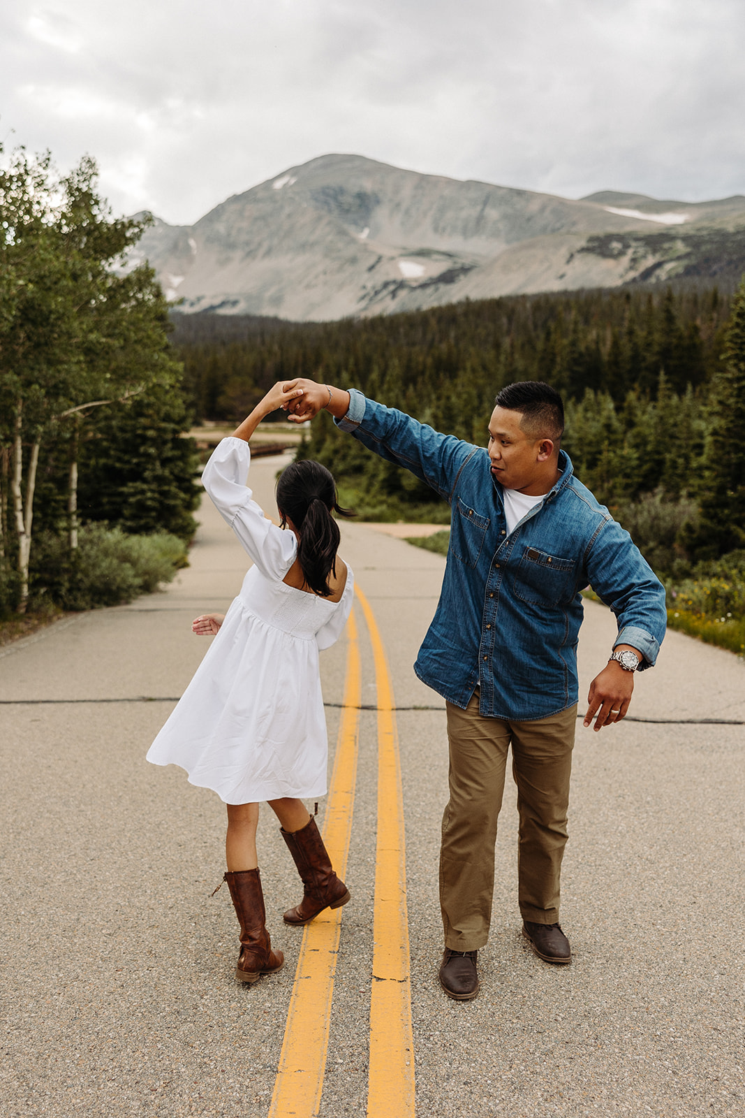 In the middle of the mountain road, the man holds his fiance's hand as she twirls under him in delight.