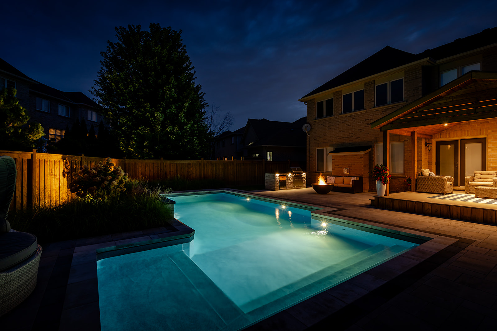 An inground pool with a fireplace on the far side of the backyard with lights on.