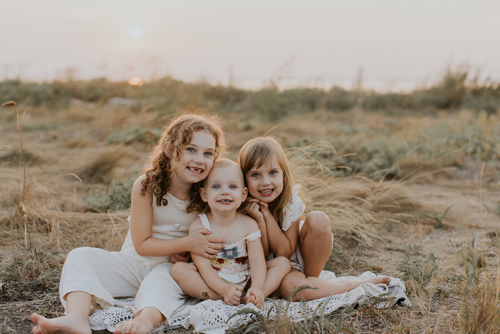 three girl siblings sitting on a blanket near the beach smiling 