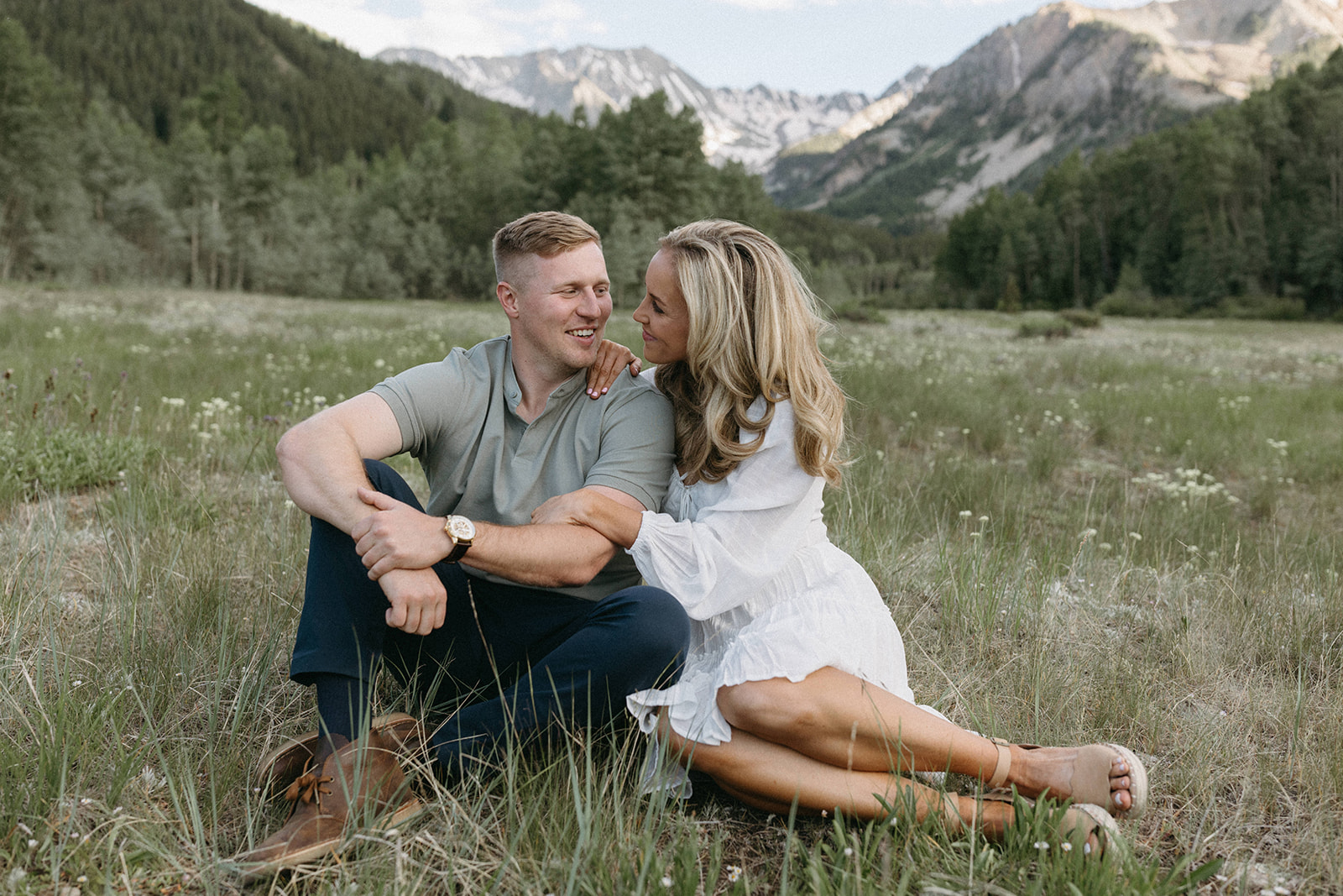 An engaged couple wearing neutral, earthy-toned engagement outfits sit in a wildflower meadow in Aspen. 