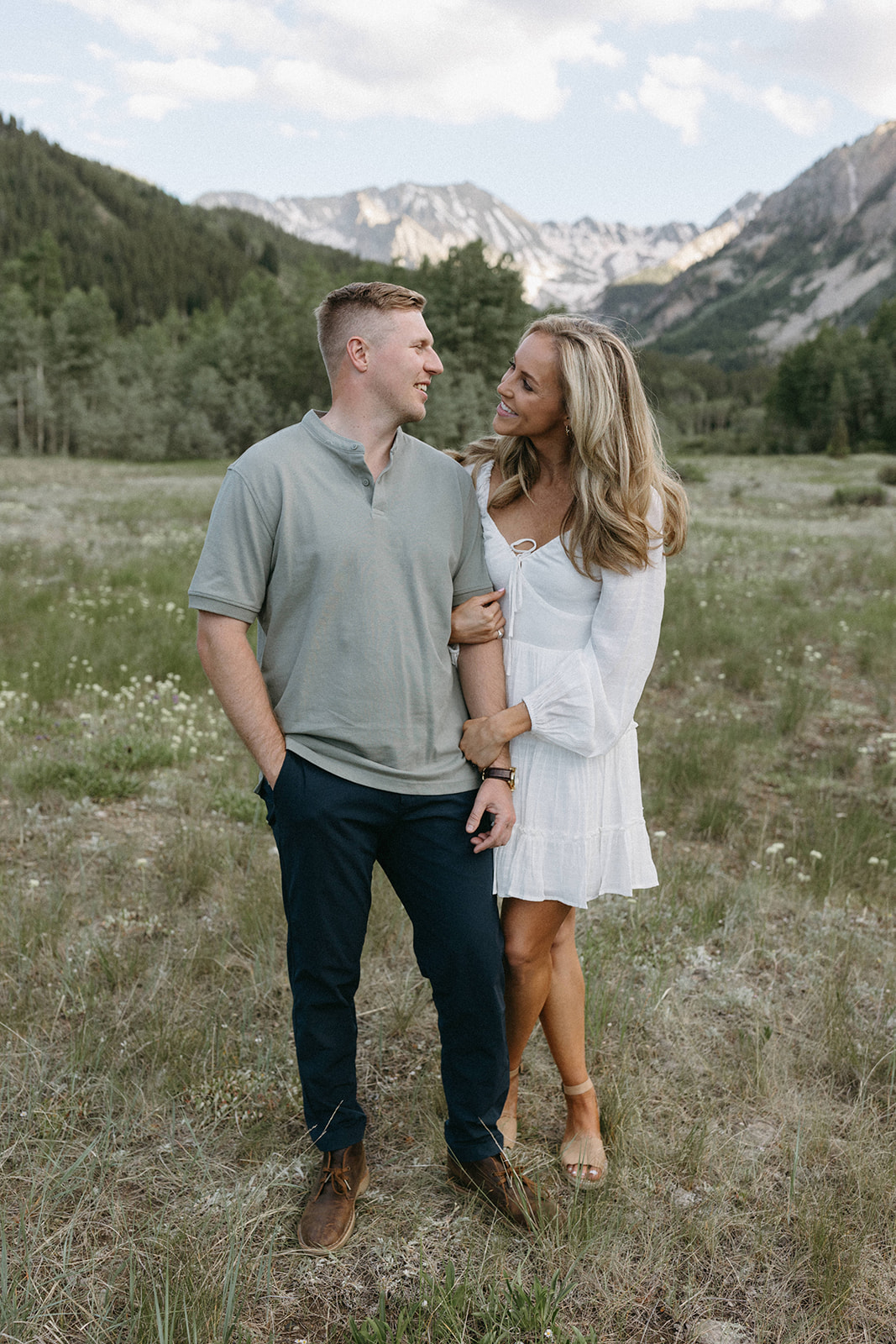 A woman in a white boho mini dress holds her fiancé's arm during their Aspen engagement session in the mountains. 