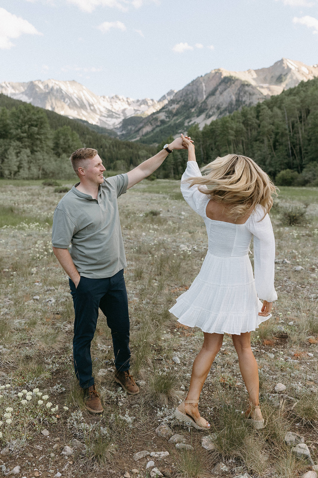 A man wearing a forest green polo shirt, chino pants, and brown chukka boots twirls a woman in a white boho dress. 