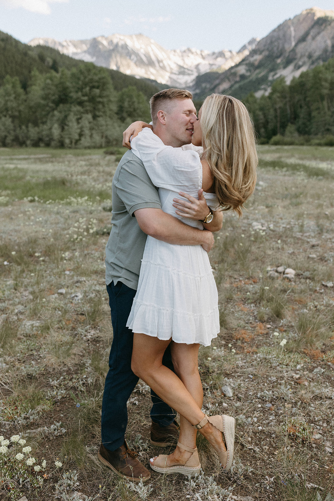 A woman in a white boho engagement dress and nude heels wraps her arms around her fiancé in an Aspen wildflower meadow. 