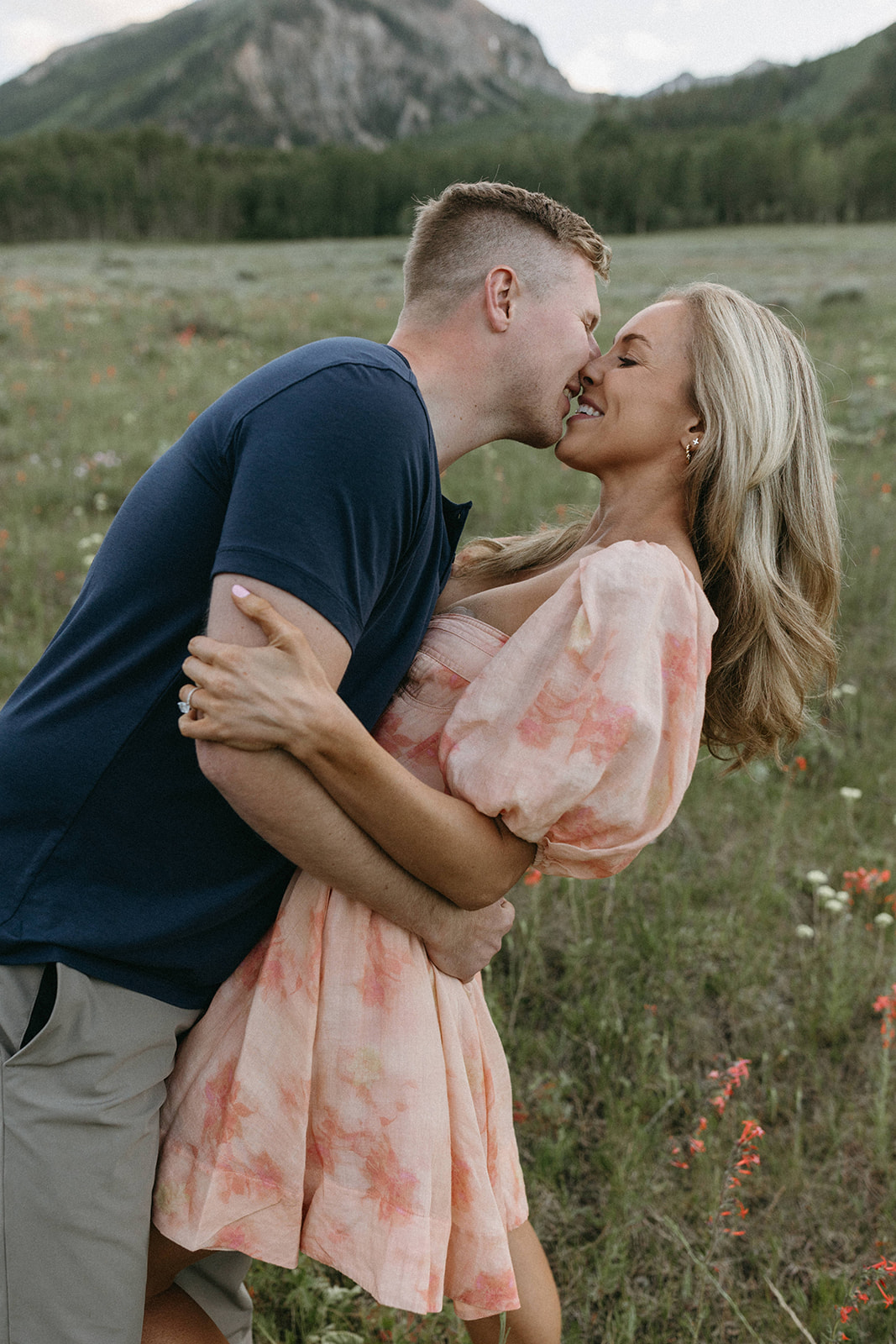 An engaged couple shares a kiss in a wildflower meadow near mountains during their Aspen engagement session in Colorado.