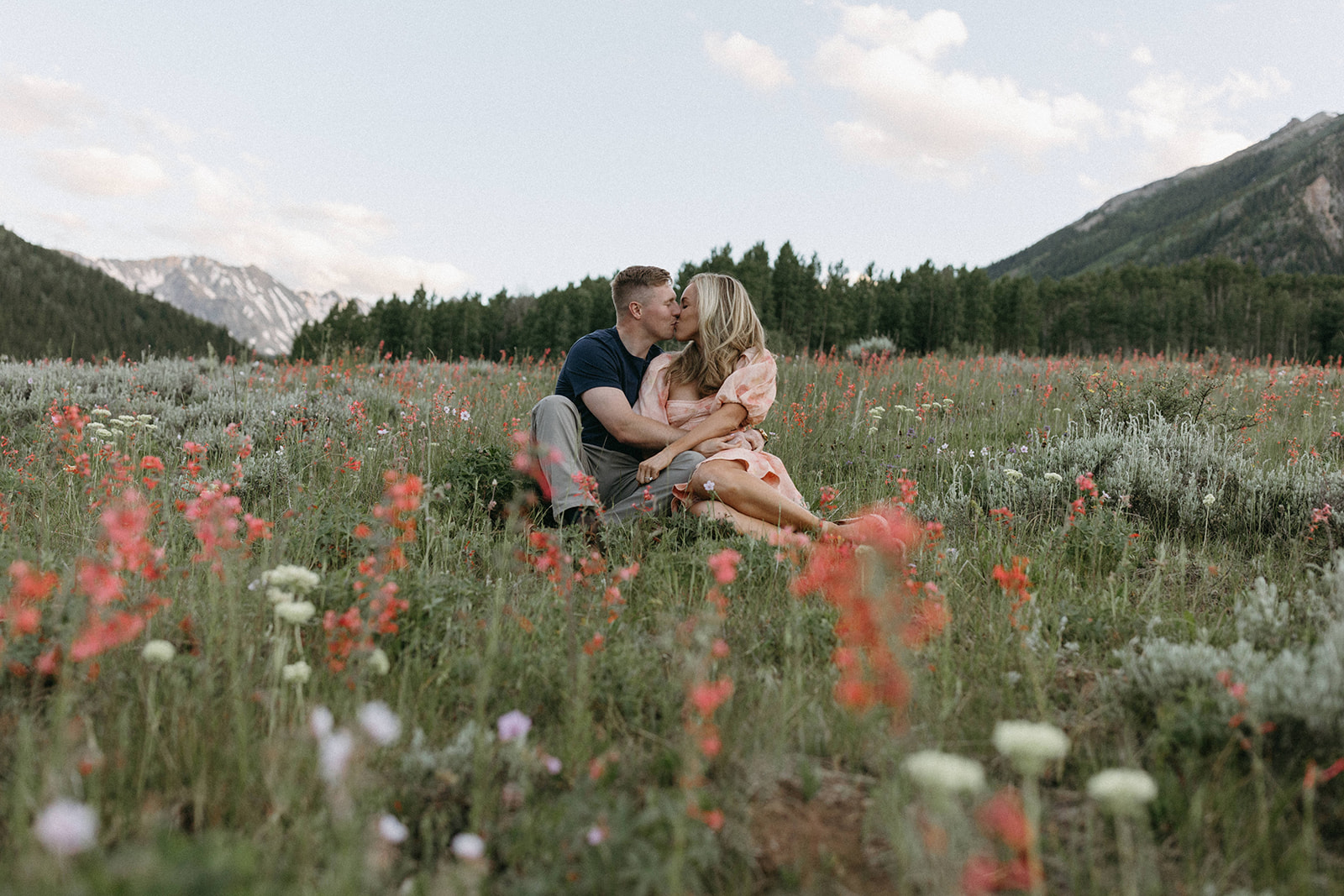 An engaged couple is seated in a wildflower meadow while sharing a kiss during their Aspen engagement session. 