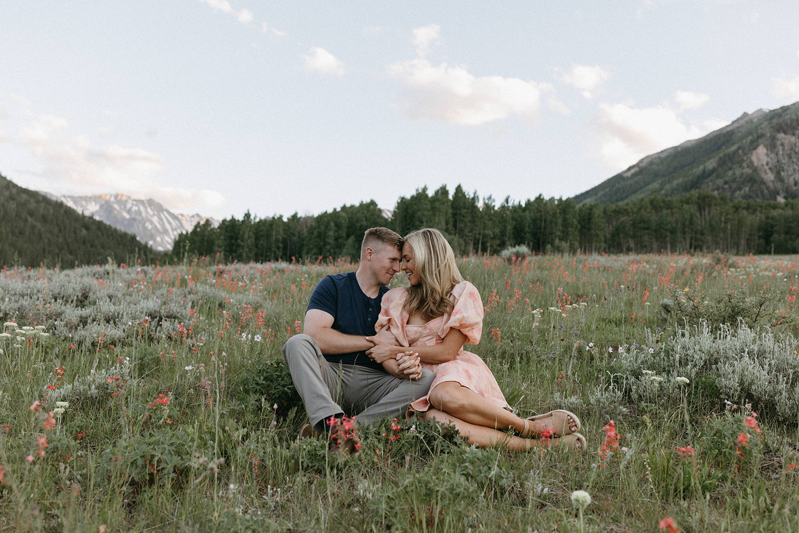 An engaged couple is seated in a wildflower meadow while getting photos taken by an Aspen Wedding Photographer. 