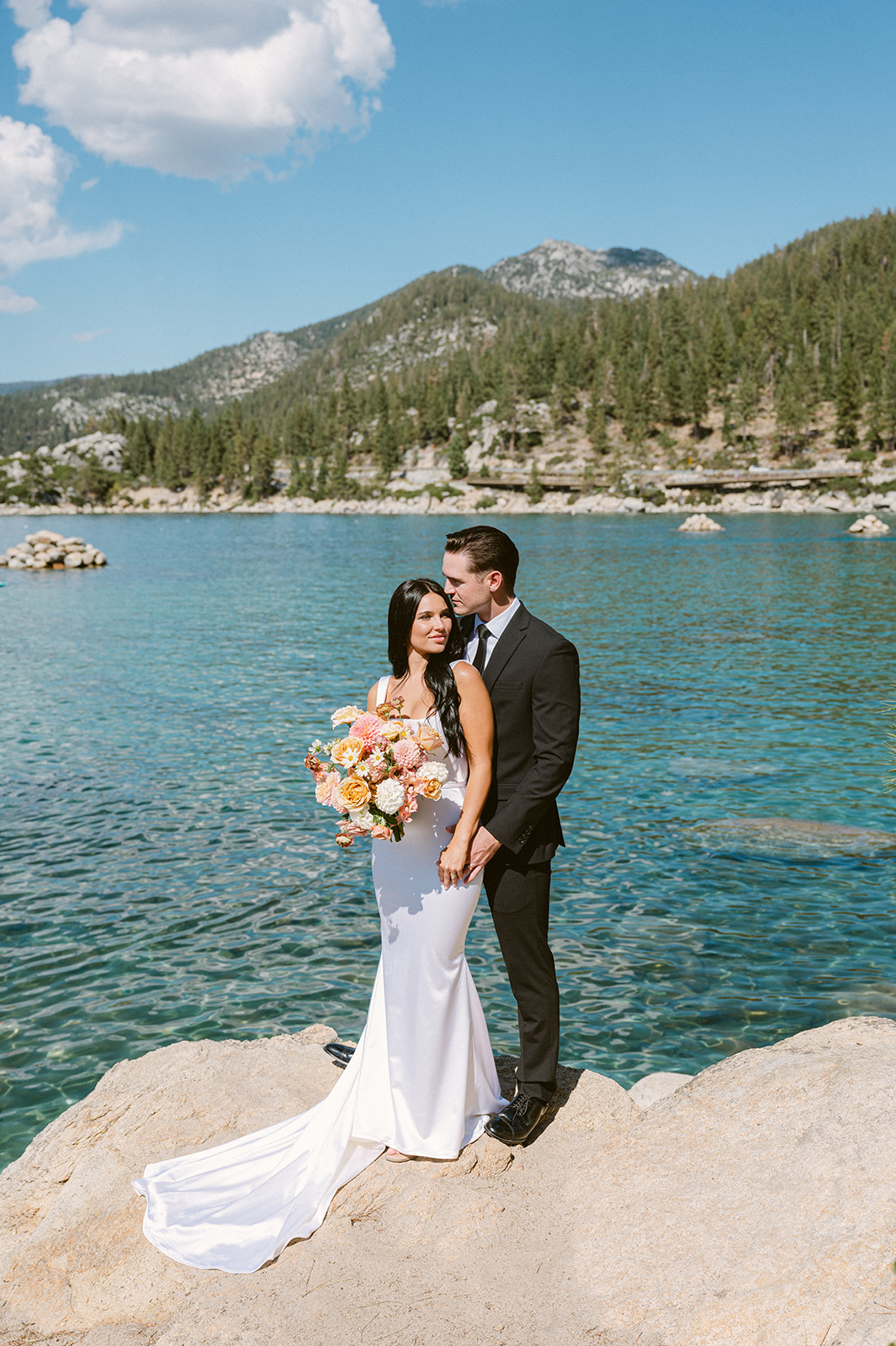bride and groom standing on large white boulders in lake tahoe with crystal blue teal water below