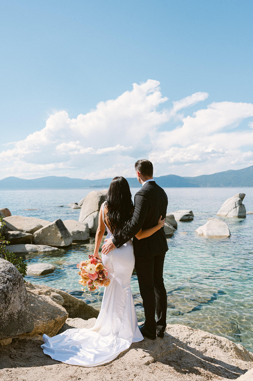bride and groom standing on large white boulders in lake tahoe with crystal blue teal water below