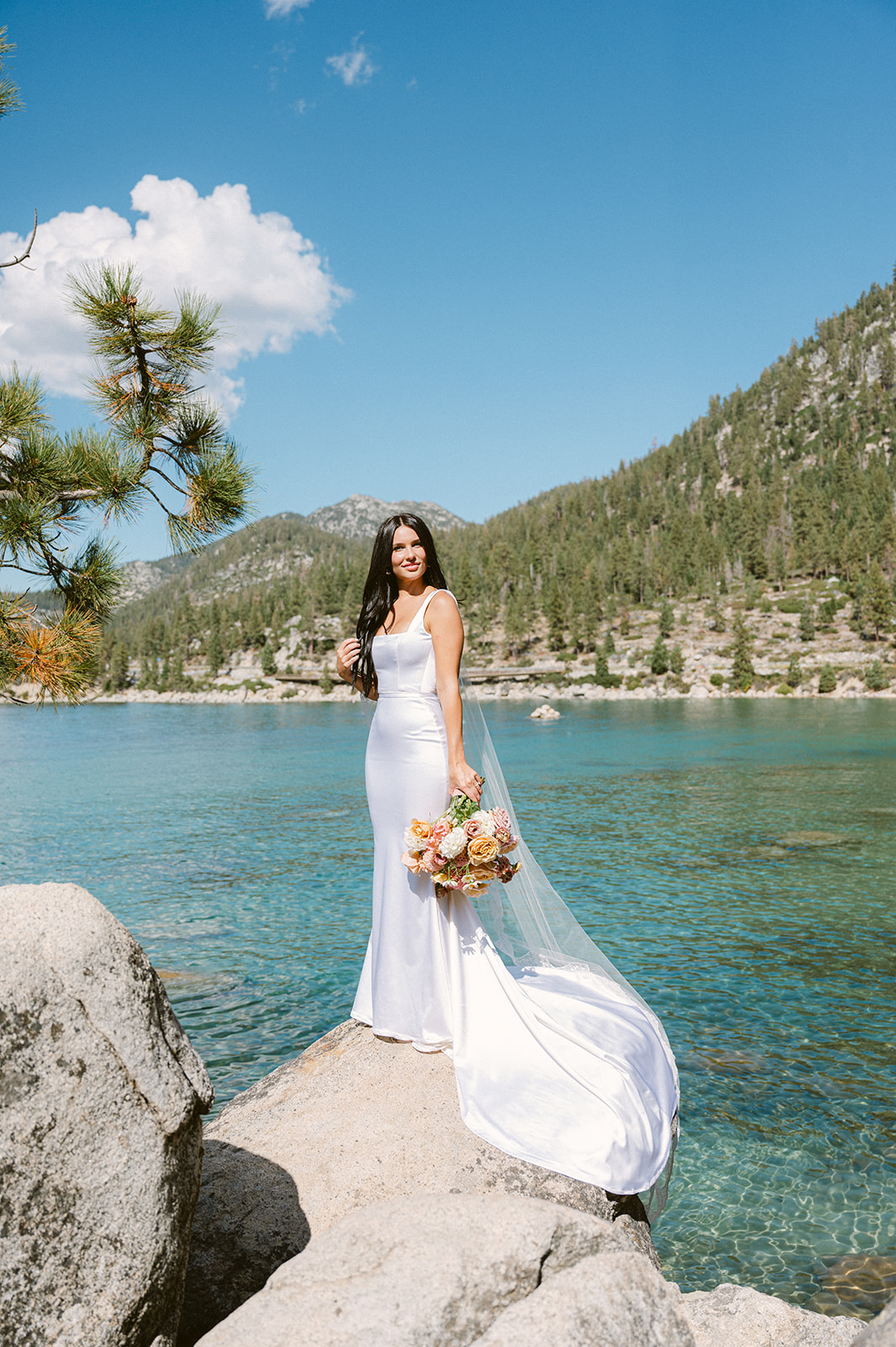 bride standing on large white boulders in lake tahoe with crystal blue teal water below